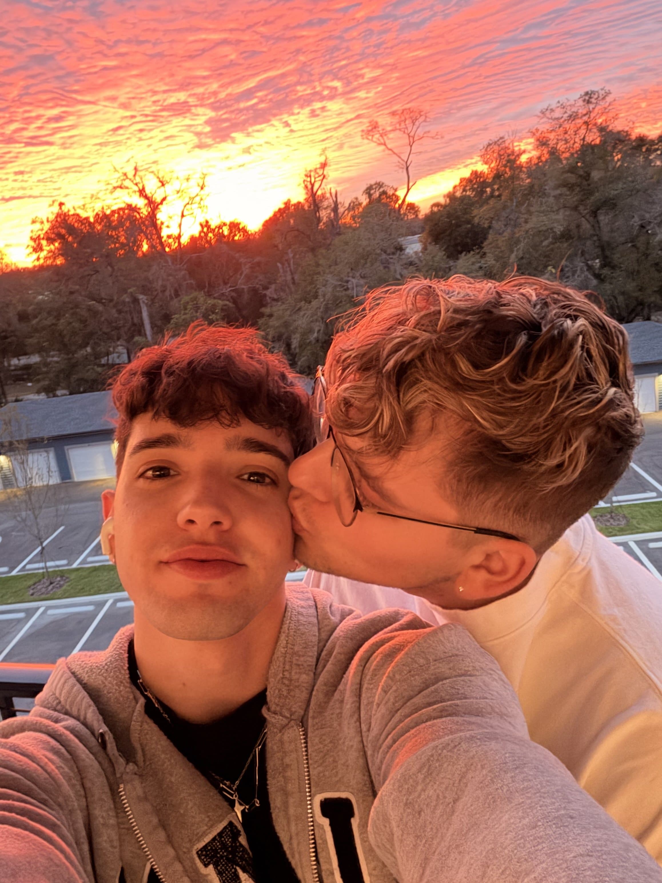 Corey kissing Damian on the cheek at golden hour against a fiery sunset sky